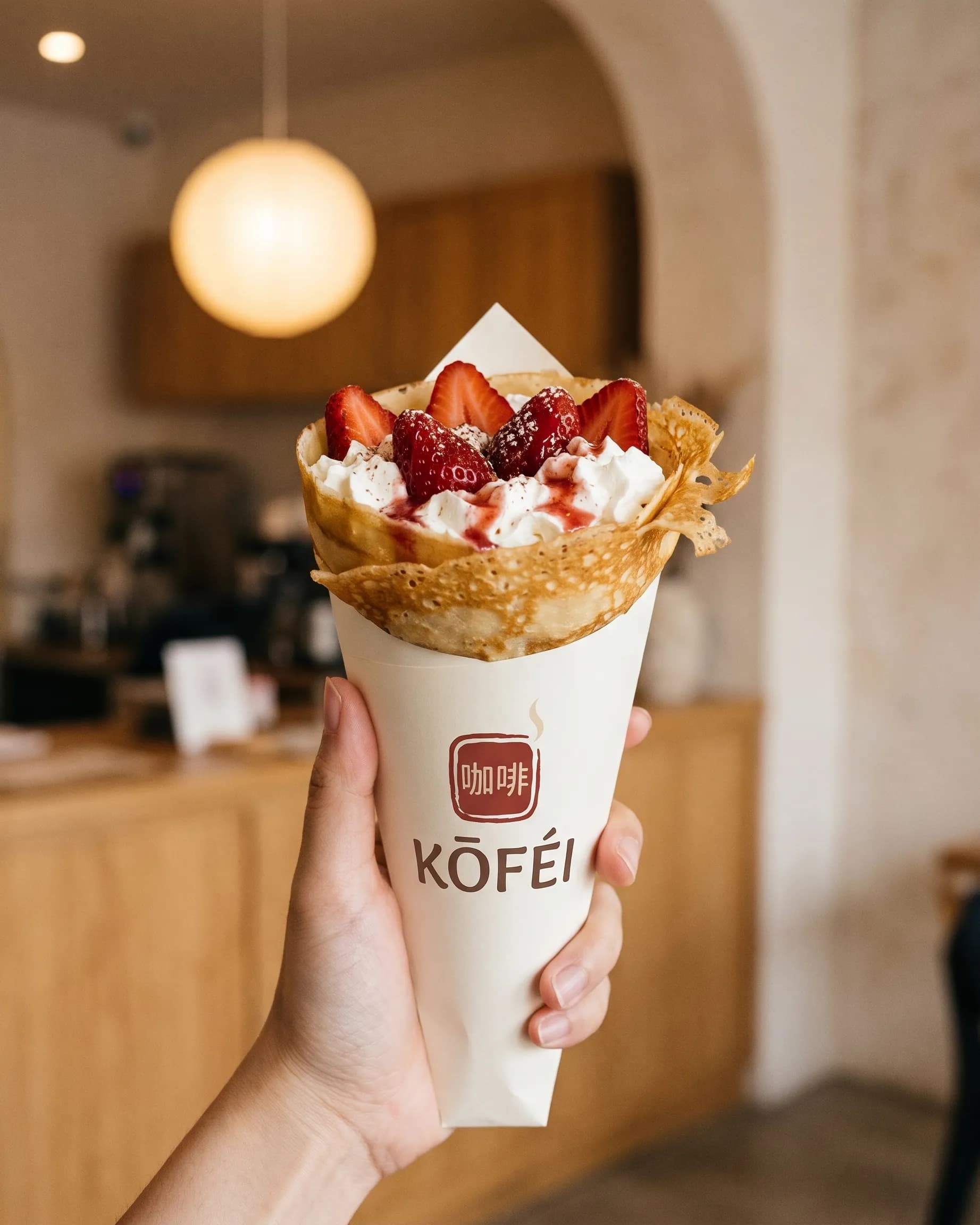 A hand holding up a strawberry and whipped-cream crêpe in a KŌFÉI paper cone, a paper lantern glowing behind